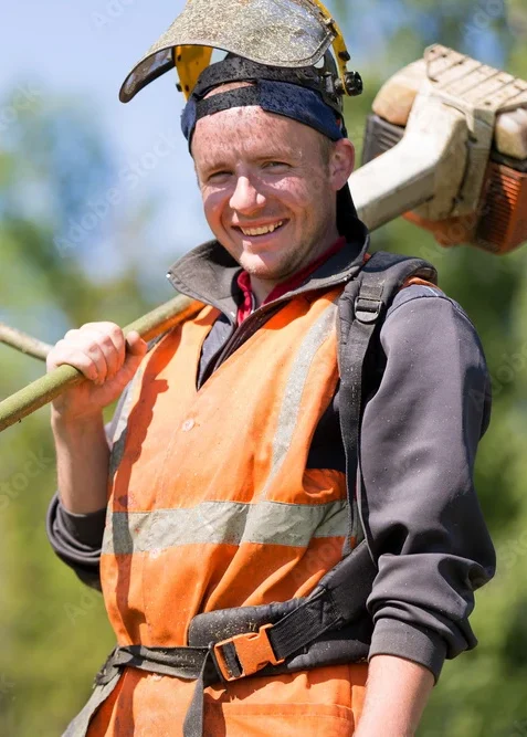 Smiling worker in an orange safety vest carrying landscaping equipment over his shoulder.