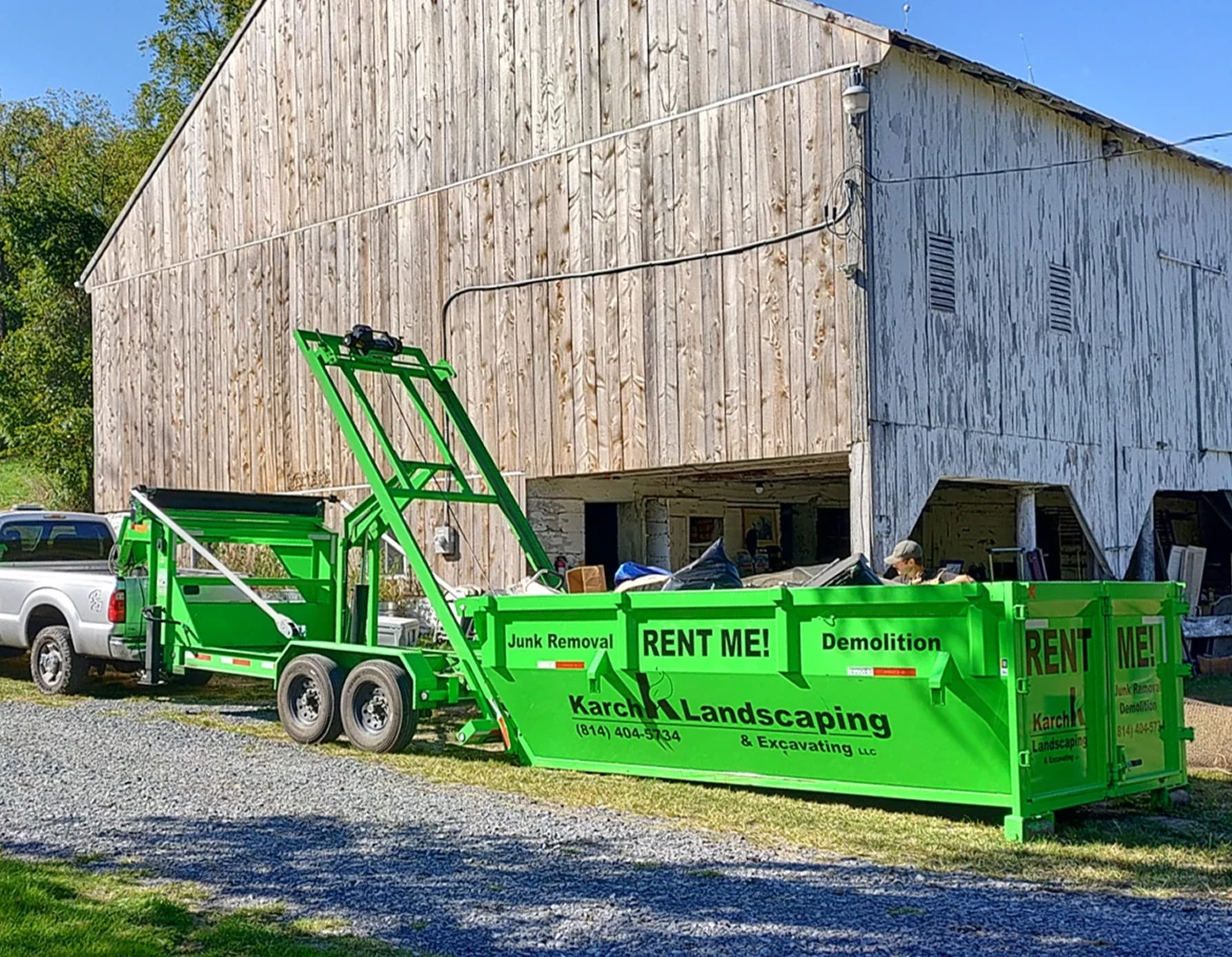 Green junk removal trailer parked next to a rustic wooden barn.