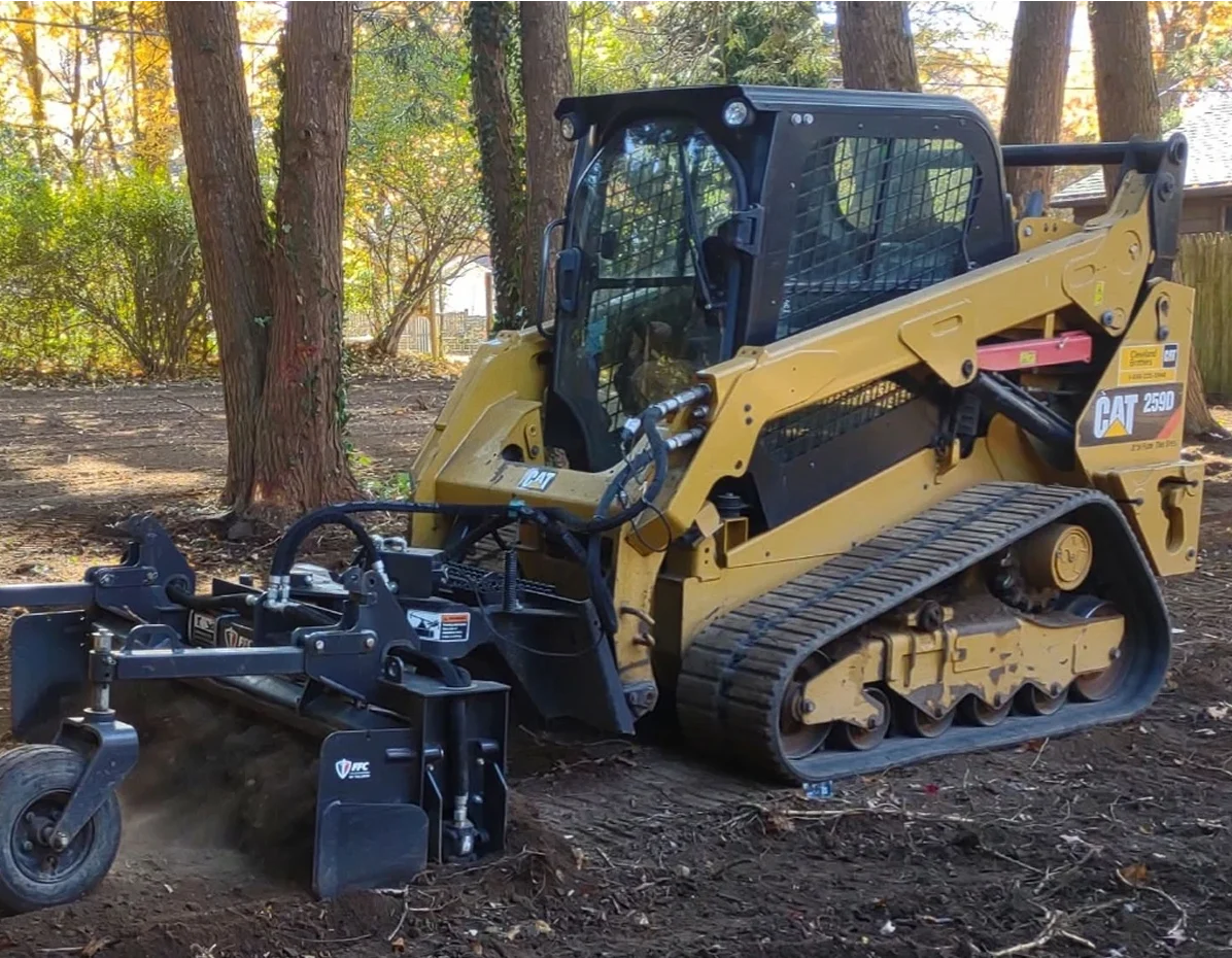 Skid steer loader with a clearing attachment working in a wooded area.