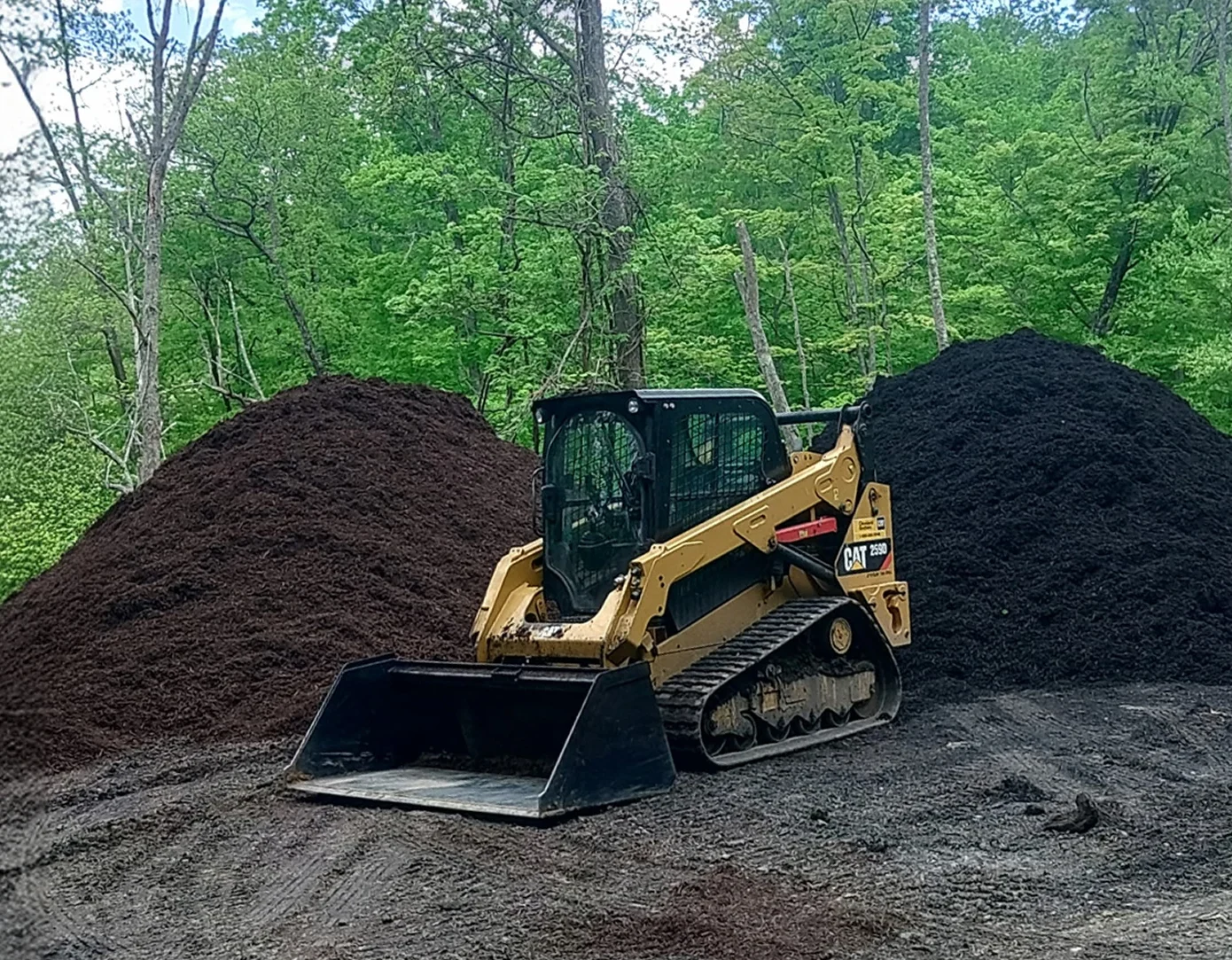 Small yellow bulldozer parked next to large mounds of dark soil or mulch.