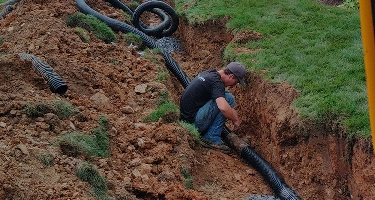 A worker installing drainage pipes in a freshly dug trench in a yard.