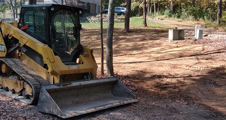 Small yellow excavator operating in a dirt area surrounded by trees.